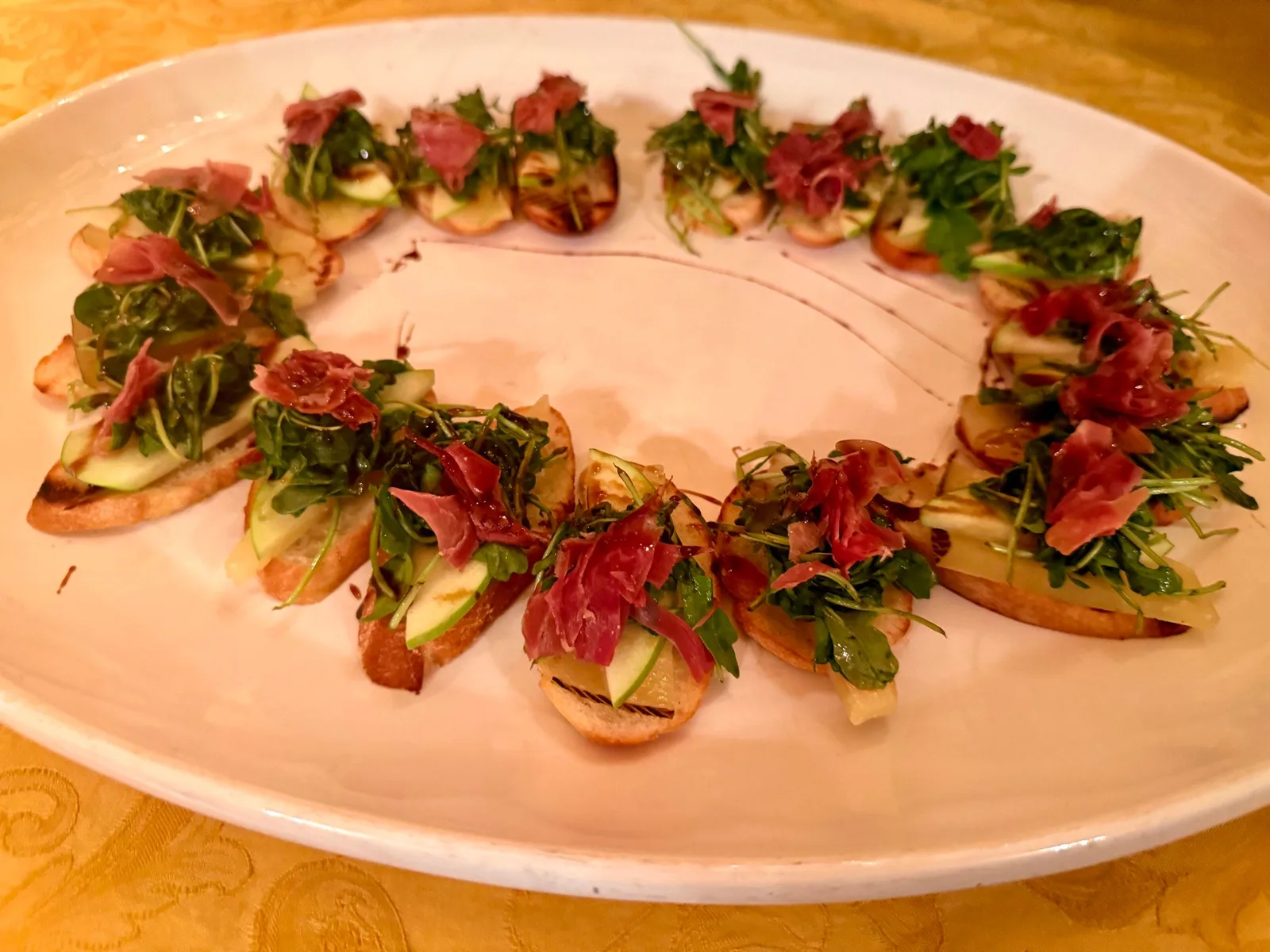 Prosciutto and arugula crostini arranged in a circle on a white oval platter